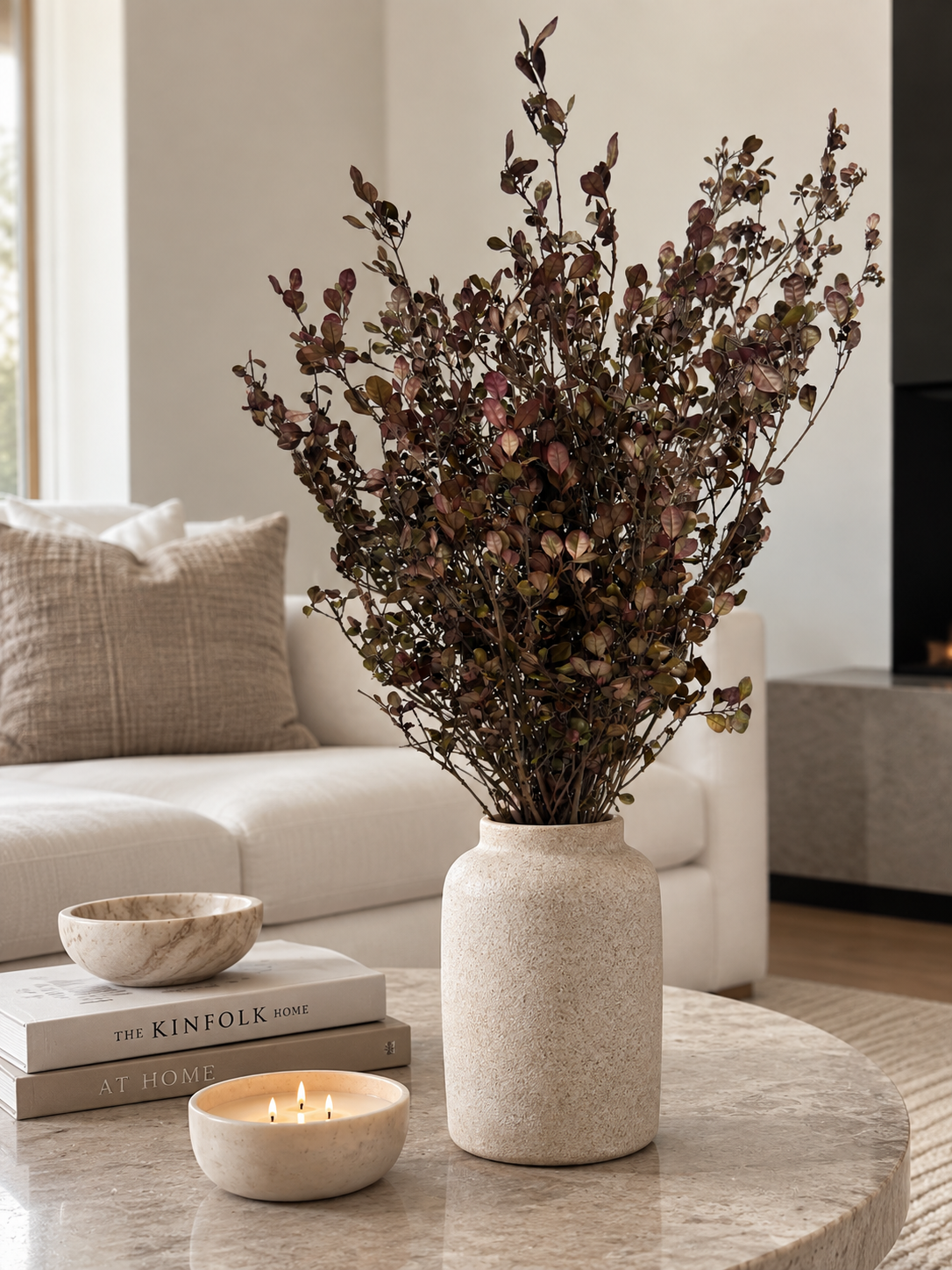 A textured cream ceramic vase filled with dried burgundy and brown foliage, styled on a marble coffee table in a modern living room setting next to a lit candle and books.