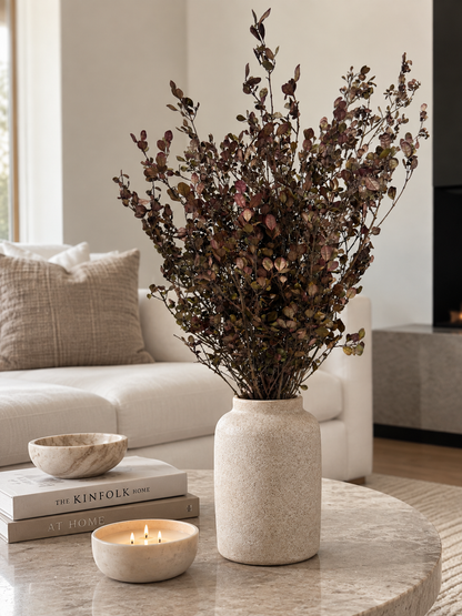 A textured cream ceramic vase filled with dried burgundy and brown foliage, styled on a marble coffee table in a modern living room setting next to a lit candle and books.